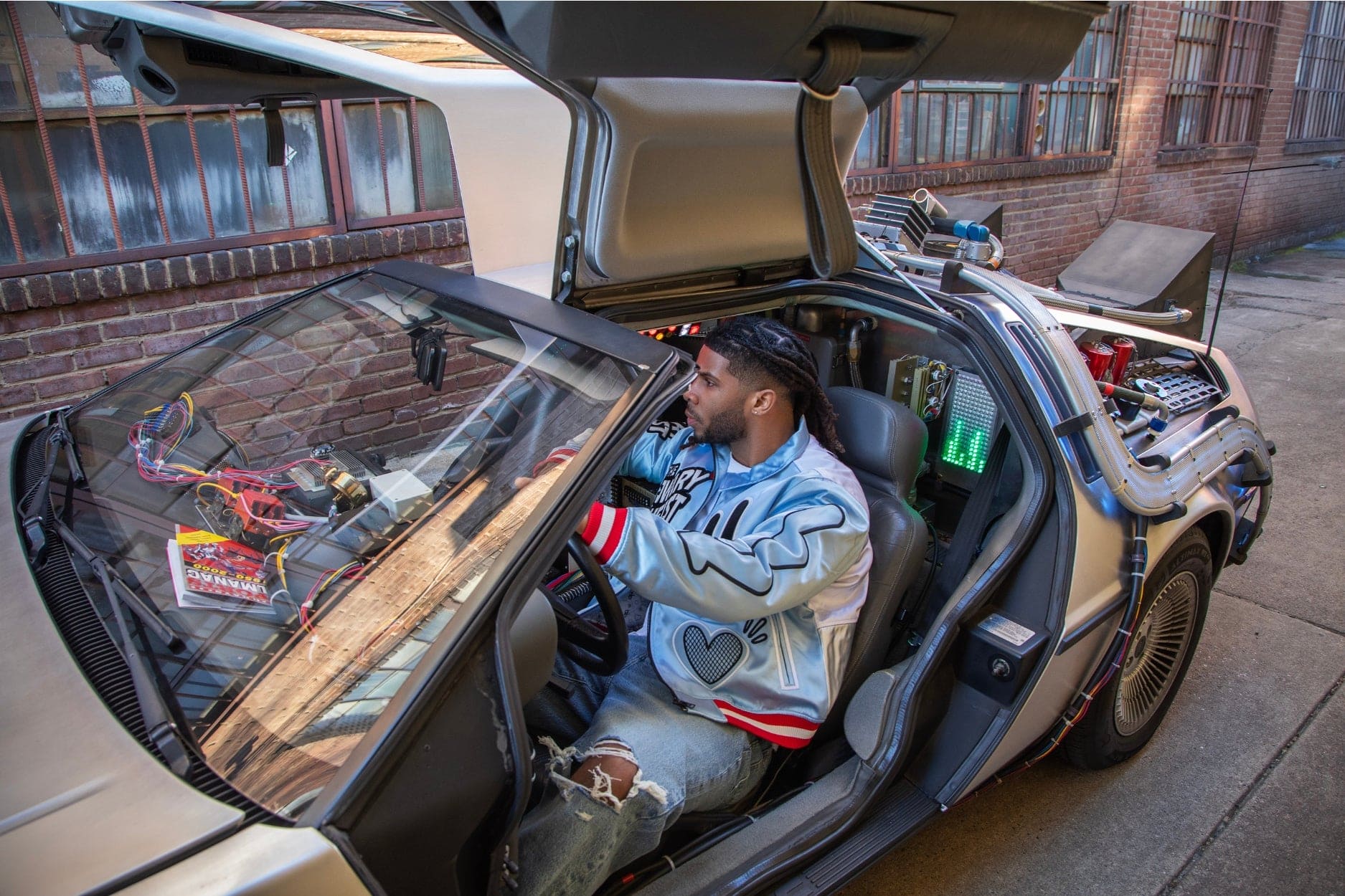 A person sitting in a modified time machine, featuring a futuristic dashboard and open doors, against a brick wall background.